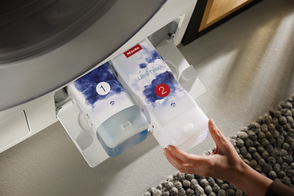 Two bottles of Miele's TwinDos solution being loaded at the bottom of washing machine.